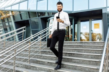 Genç yakışıklı işadamı cep telefonu kullanıyor ve ofis binasının yanındaki sokakta duruyor.