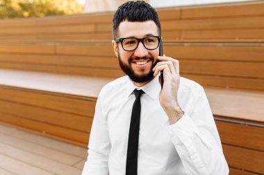 Genç yakışıklı iş adamı gözlüklü, bankta oturuyor ve telefonda konuşuyor.