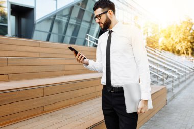 Genç yakışıklı işadamı cep telefonu kullanıyor ve ofis binasının yanındaki sokakta duruyor.