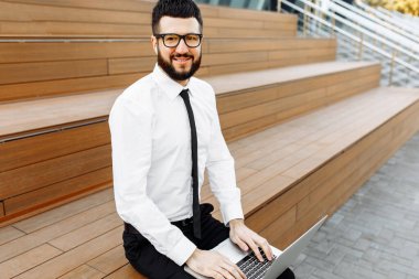 Gözlüklü şık bir işadamı laptopla çalışıyor, merdivenlerde kahveyle oturuyor.