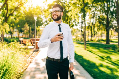 Bardaklı mutlu bir iş adamı, elinde bir fincan kahveyle, sokakta parkta yürüyor.