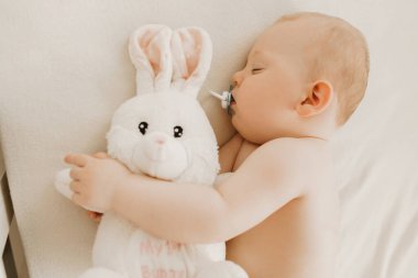 Küçük yeni doğmuş bebek beşikte pelüş beyaz bir tavşanla uyuyor.