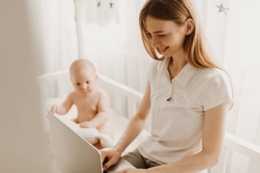 Güzel, genç bir anne dizüstü bilgisayarla çalışıyor ve evde küçük bir çocukla oturuyor. Uzaktan çalışma, karantina...
