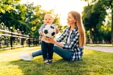 Parktaki çimlerin üzerinde annesinin futbol topuyla oynadığı neşeli bir çocuk.