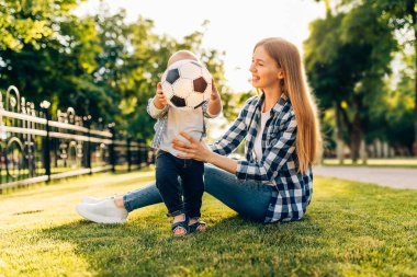 Parktaki çimlerin üzerinde annesinin futbol topuyla oynadığı neşeli bir çocuk.