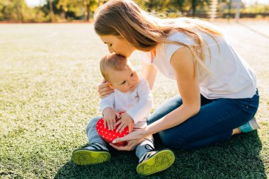 Mutlu küçük çocuk gülümseyen annesini tebrik eder ve parkta çimenlerde otururken ona kırmızı bir kalp hediye eder, Anneler Günü.