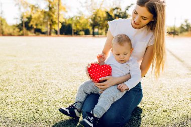 Mutlu küçük çocuk gülümseyen annesini tebrik eder ve parkta çimenlerde otururken ona kırmızı bir kalp hediye eder, Anneler Günü.