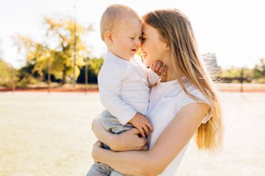 Bebeğiyle ilgilenen genç bir anne, sevgi dolu bir anne küçük çocuğu kucaklıyor güneşli bir günde dışarıda şefkatli bir aile anının tadını çıkarıyor.