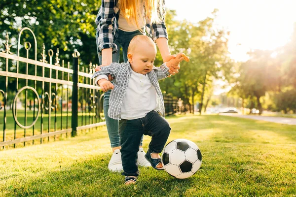 Happy young mom and her little son play soccer together outdoors in the park