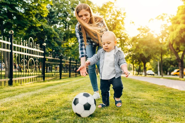 Happy young mom and her little son play soccer together outdoors in the park