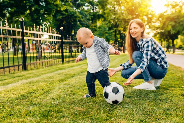 Happy young mom and her little son play soccer together outdoors in the park