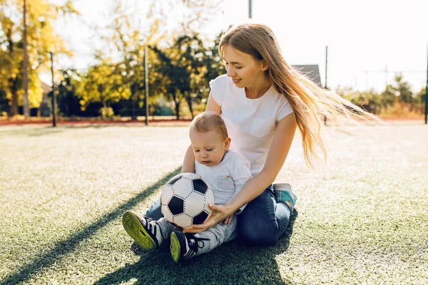 Happy young mom with son playing with soccer ball on field, outdoors