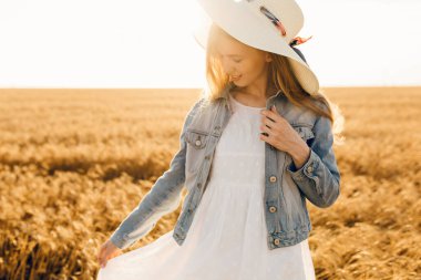 Gün batımında buğday tarlasında giysili ve şapkalı mutlu bir genç kadın.