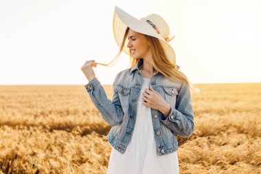Güzel çiftçi kız buğday tarlasında şapka takıyor ve günbatımının tadını çıkarıyor.
