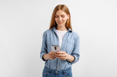 Neşeli genç kadın beyaz arkaplandaki akıllı telefon ekranına bakarak SMS mesajı yazıyor.