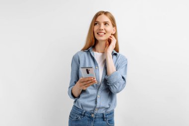 Düşünceli genç bir kadın cep telefonu kullanıyor ve beyaz arkaplanda fotokopi uzayına bakıyor.