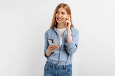 Düşünceli genç bir kadın cep telefonu kullanıyor ve beyaz arkaplanda fotokopi uzayına bakıyor.