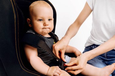 Anne küçük bebeğini bebek koltuğuna bağlıyor, bebek de bebek koltuğuna, izole edilmiş beyaz bir arka plana.