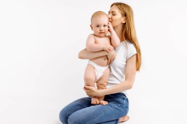 Genç anne ve bir yaşındaki oğlunun portresi, sevgi dolu anne bebeğine sarılıyor ve onu beyaz arka planda öpüyor.