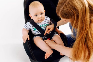 Anne küçük bebeğini bebek koltuğuna bağlıyor, bebek de bebek koltuğuna, izole edilmiş beyaz bir arka plana.