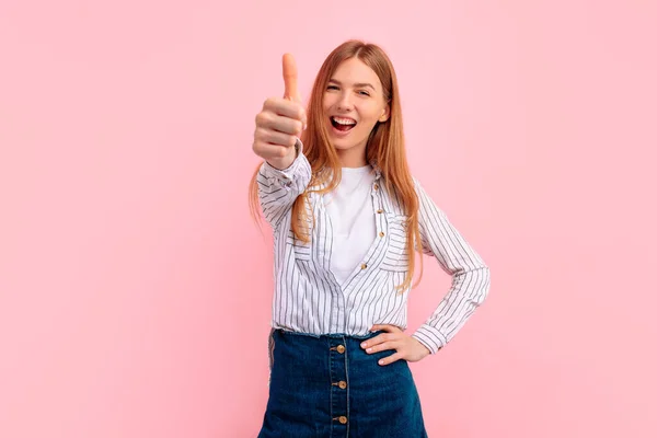 Happy young woman making a thumb up sign and smiling, showing her support and respect, on a pink background, I like it