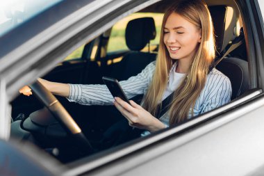 Güneş gözlüklü genç bir kadın araba sürerken cep telefonu kullanıyor.