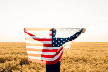 Çekici bir adam açık güneşli bir günde mavi bir gökyüzüne karşı Birleşik Devletler bayrağını tutuyor, arkadan bakıyor, yakın plan. Ulusal tatil konsepti