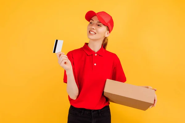 快乐的年轻女子信使 身穿红色制服 送货员手持纸盒和信用卡 背景是孤立的黄色 — 图库照片
