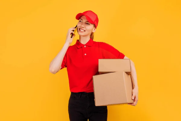 送货女工头戴红帽子 身穿T恤 手持纸板箱 手持手机 背景是黄色的 — 图库照片