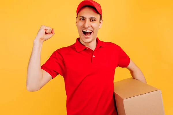 身穿红帽子和T恤 手持纸板箱 打着黄色背景庆祝胜利的快乐送货员 — 图库照片
