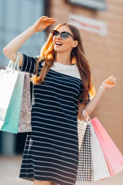 Heyecanlı, şatafatlı, güneş gözlüklü, renkli alışveriş çantaları olan alışveriş merkezinin arka planında caddede duran mutlu, şık bir kadın. Alışveriş, moda.