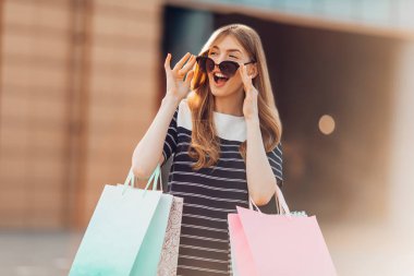 Heyecanlı, şatafatlı, güneş gözlüklü, renkli alışveriş çantaları olan alışveriş merkezinin arka planında caddede duran mutlu, şık bir kadın. Alışveriş, moda.