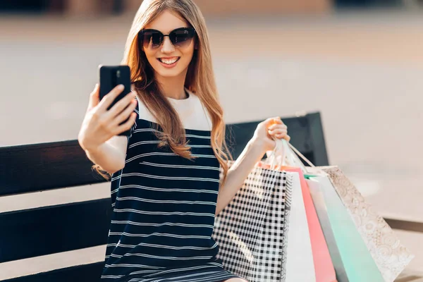 선글라스를 매력적 아름다운 쇼핑백을 쇼핑센터 벤치에 전화로 셀카를 온라인 — 스톡 사진