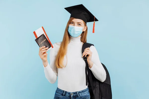 Happy young woman student in medical mask, student in graduation hat holding passport and tickets on blue background, Study, education, university, admission