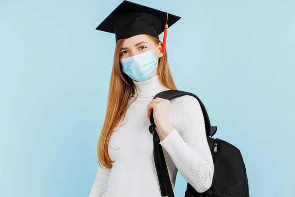 Happy young woman graduate in medical mask, student in graduation hat with briefcase on blue background