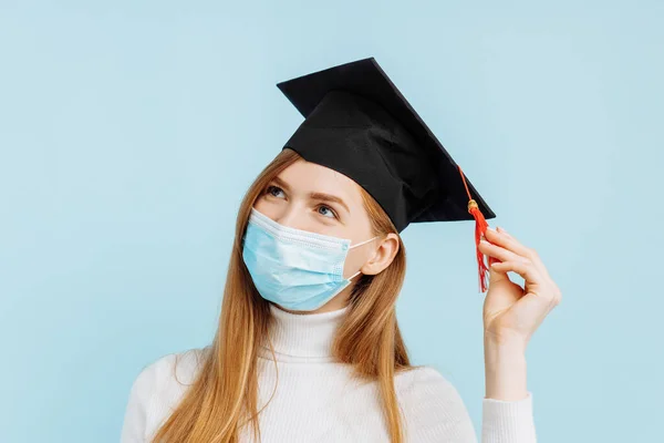Happy thinking graduate in medical mask, student in graduation hat with briefcase thinks and looks to the side on blue background