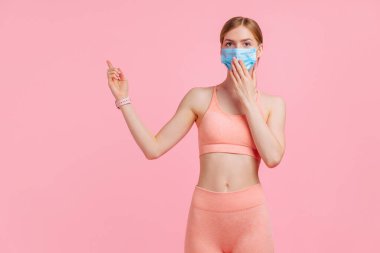 Yüzünde tıbbi koruyucu maske takan, spor kıyafetli güzel genç bir kadın pembe arka planda fotokopi alanının kenarına bakıyor ve işaret ediyor.