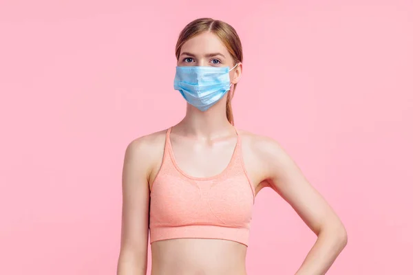 Beautiful young fitness woman wears protective mask to prevent corona virus, sportswoman with perfect body wears sportswear for workouts, on isolated pink background