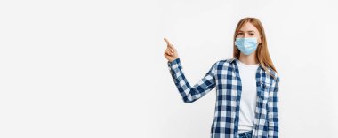 Tıbbi koruyucu maskeli genç bir kadın izole edilmiş beyaz arka planda fotokopi uzayını işaret ediyor.