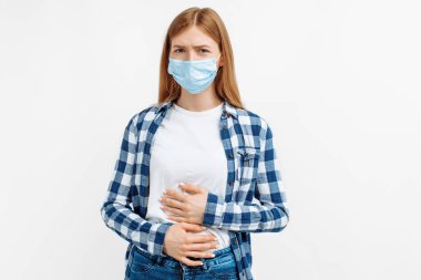 Korumacı maskeli genç bir kadın iki elini karnının üstüne koyarak izole edilmiş beyaz arka planda karın ağrısı çekiyor.