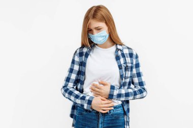 Korumacı maskeli genç bir kadın iki elini karnının üstüne koyarak izole edilmiş beyaz arka planda karın ağrısı çekiyor.