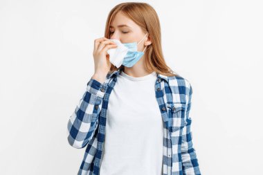 Hasta genç bir kadın, koruyucu bir maske takıyor, burnunu siliyor, ateşi var, nezle oluyor, izole edilmiş beyaz bir arka planda hapşırıyor.