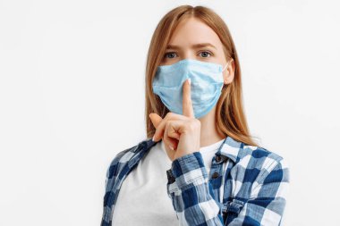 Virüslerden korunan koruyucu maskeli genç bir kadın, işaret parmağını dudaklarının üstünde tutuyor, sessizlik jesti yapıyor, sessiz olmak istiyor, izole edilmiş beyaz bir arka planda.