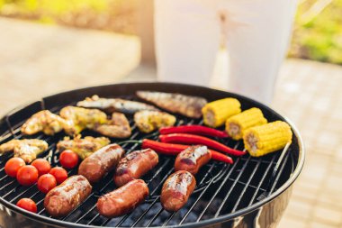 Barbekü yapan bir adam, çeşitli sebzeler ve sosisli tavuk kanatları, doğadaki bir parkta portatif bir barbeküde ızgara yapan bir adam.