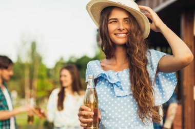 Yazlık şapkalı ve güneş gözlüklü güzel bir kadın, arka planda bir grup arkadaşla dışarıda barbekü yapıyorlar bira içiyorlar, güneşli bir günde dışarıda gülüyorlar, yaz zamanı, piknik yapıyorlar.
