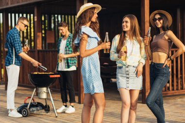 Bir grup genç öğrenci terasta eğleniyor. Bira içip sohbet ediyorlar. Arkadaşları da yaz tatilinde mangalda et servis ediyor.