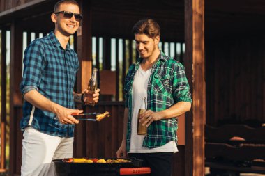 Bira içen iki genç adam, doğada barbekü yapıyorlar, şık insanlar et, gençlik yaşam tarzı, yemek, arkadaşlık ve yaz konsepti hazırlıyorlar.