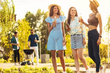 Üç güzel kız arkadaş piknikte eğleniyor, içki içiyor ve sohbet ediyor, bir grup genç arkadaş gün batımında parkta ızgarada barbekü yapıyor.