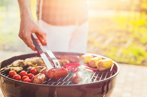 男はグリルで肉や野菜をフライドポテト トングとソーセージを反転 トウモロコシのコブ 夏に屋外唐辛子と鶏の羽 — ストック写真