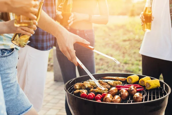 Barbekü yapan bir adam, çeşitli sebzeler ve sosisli tavuk kanatları, doğadaki bir parkta portatif bir barbeküde ızgara yapan bir adam.